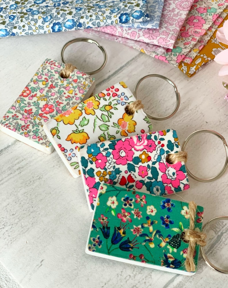 May include: Four keychains with different floral patterns. Each keychain has a silver ring and a brown twine loop. The patterns include a white background with pink, yellow, and blue flowers, a white background with pink and blue flowers, a white background with yellow and blue flowers, and a green background with blue, pink, and white flowers.