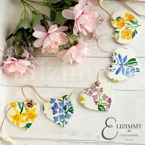 Puede incluir: Una guirnalda de cinco adornos blancos en forma de corazón con motivos florales, incluyendo abejas, mariposas y libélulas. La guirnalda está enhebrada con un cordel y cuelga sobre un fondo de madera blanca.