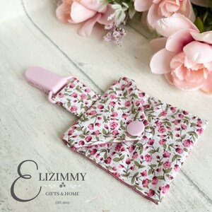 Puede incluir: Babero de bebé de tela floral rosa con un clip de plástico rosa. El babero tiene un pequeño bolsillo y un botón rosa. La tela tiene un fondo blanco con pequeñas flores rosas y hojas verdes. El texto "Lizimmy Gifts & Home Est. 2012" está impreso en la parte inferior del babero.