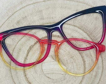 May include: Two pairs of eyeglass frames. The top pair features a dark blue to magenta gradient with a cat-eye shape. The bottom pair has a rounded shape with a vibrant gradient transitioning from a deep red at the top to a bright yellow at the bottom.