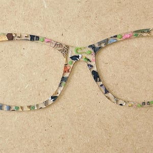May include: Cat-eye spectacles with a clear frame adorned with colourful animal illustrations. The frame features various animals, including giraffes, elephants, and flamingos. The glasses are set against a neutral background.