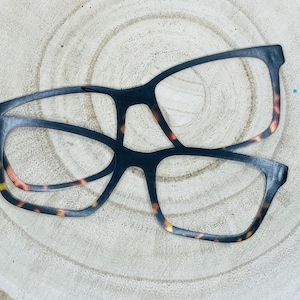 May include: Two pairs of spectacles with black frames and tortoiseshell detailing. The glasses have a rectangular shape and are placed on a light-coloured, wood-grain surface. The frames are thick, suggesting a classic design.