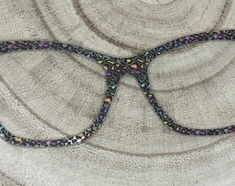 May include: A pair of eyeglasses with a cat-eye frame. The frame features a leopard print pattern in shades of black, gray, and iridescent colors. The glasses are set against a light-colored, textured background.