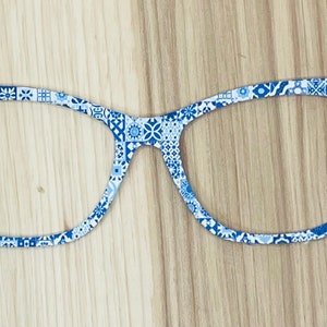 May include: A flat, two-dimensional cutout in the shape of cat-eye glasses featuring a repeating blue and white geometric tile pattern. The object has a thin frame design with no lenses, showcasing a variety of intricate square motifs across the surface.
