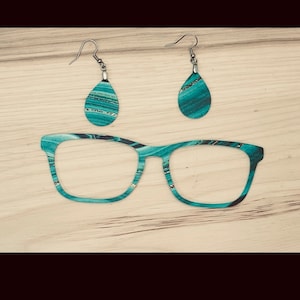 May include: Teal and gold patterned glasses and earrings. The glasses have a square frame and the earrings are teardrop shaped.