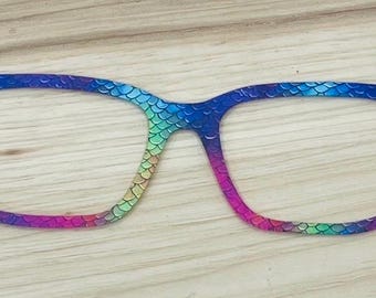 May include: A pair of eyeglass frames featuring a vibrant, multi-colored gradient pattern that transitions from green and blue to purple and pink. The surface of the frames is covered in a repeating, textured scale pattern resembling fish scales.
