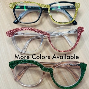 May include: Three pairs of eyeglasses with glitter frames. The frames are gold, red, and green. The text "More Colors Available" is printed below the glasses.
