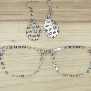 May include: Clear eyeglasses frames and two teardrop-shaped earrings, all adorned with a repeating pattern of various dog faces. The earrings have silver hooks. The items are displayed on a light wood surface.