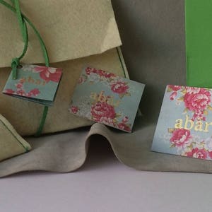 May include: A set of small, cream-colored gift bags with green ribbon ties. Each bag has a floral patterned tag attached with the word "abar" printed in gold.
