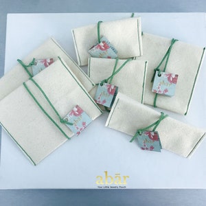 K&ouml;nnte beinhalten: Ein Set aus acht kleinen, wei&szlig;en Stoffbeuteln mit gr&uuml;nem Besatz und floralen Etiketten, auf denen "abar" steht. Die Beutel sind auf einer wei&szlig;en Oberfl&auml;che angeordnet, auf der der Text "abar Your Little Jewelry Pouch" gedruckt ist.