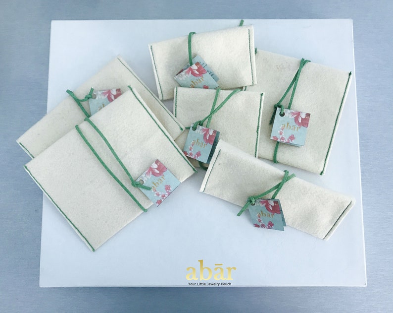 K&ouml;nnte beinhalten: Ein Set aus acht kleinen, wei&szlig;en Stoffbeuteln mit gr&uuml;nem Besatz und floralen Etiketten, auf denen "abar" steht. Die Beutel sind auf einer wei&szlig;en Oberfl&auml;che angeordnet, auf der der Text "abar Your Little Jewelry Pouch" gedruckt ist.