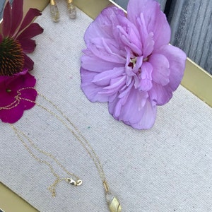 K&ouml;nnte beinhalten: Eine goldene Halskette mit einem Muschelanh&auml;nger auf einer wei&szlig;en Stoffoberfl&auml;che mit einer lila Blume und goldenen Ohrringen.
