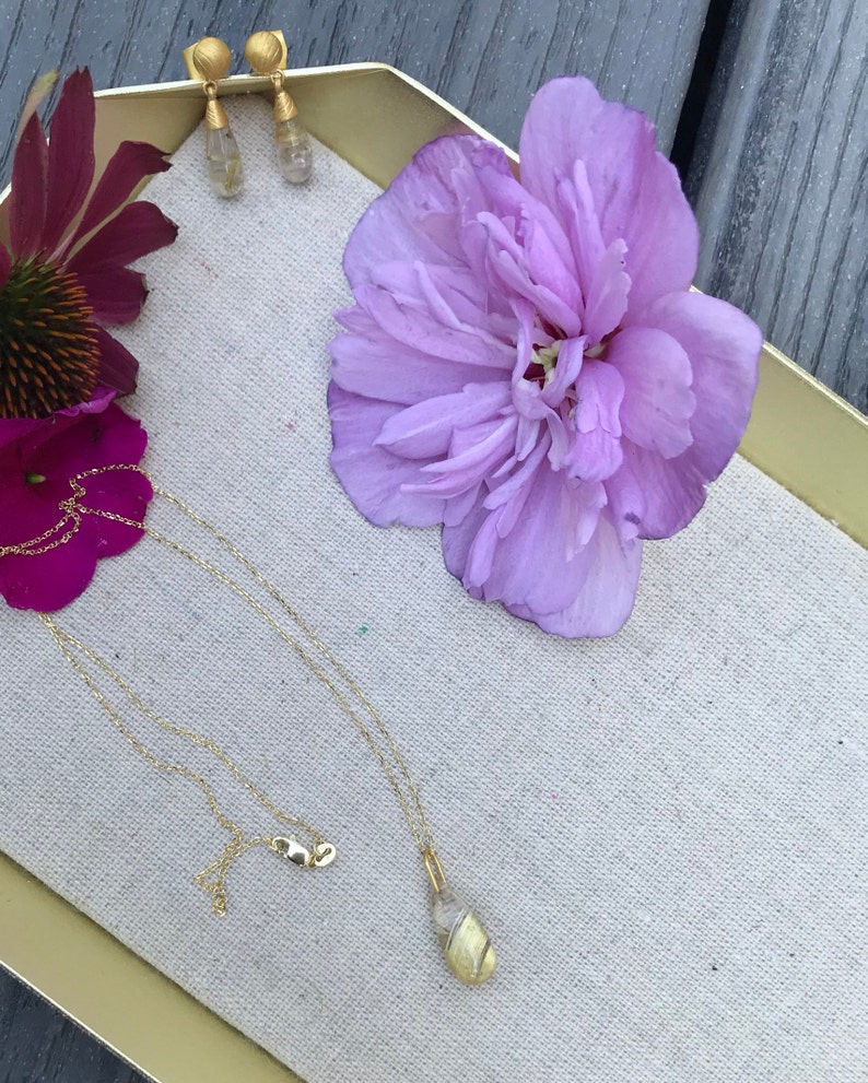 K&ouml;nnte beinhalten: Eine goldene Halskette mit einem Muschelanh&auml;nger auf einer wei&szlig;en Stoffoberfl&auml;che mit einer lila Blume und goldenen Ohrringen.