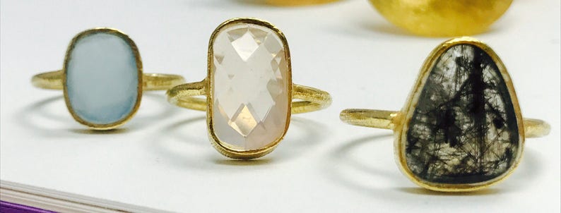 May include: Three gold rings with different gemstone settings. The first ring has a light blue oval-shaped gemstone. The second ring has a clear oval-shaped gemstone. The third ring has a dark gray, almost black, oval-shaped gemstone.