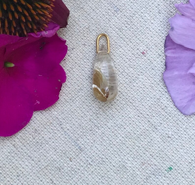 K&ouml;nnte beinhalten: Ein tropfenf&ouml;rmiger Anh&auml;nger mit einer goldenen &Ouml;se und einem klaren Harz, gef&uuml;llt mit getrockneten Blumen.