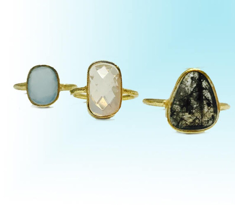 May include: Three gold rings with different colored gemstones. The first ring has a light blue oval-shaped stone. The second ring has a clear, rectangular stone. The third ring has a dark gray, triangular stone.