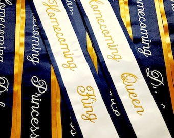May include: A close-up of several white sashes with gold lettering. The sashes are for a homecoming event and read "Homecoming King", "Homecoming Queen", "Homecoming Princess", and "Homecoming". The sashes are decorated with gold and navy blue ribbon.