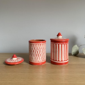 May include: Two red ceramic jars with white geometric patterns. One jar has a lid with a star design. The jars are on a wooden surface.