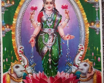 May include: A colorful print depicting a Hindu goddess, adorned in green and red garments, holding lotus flowers. The image features ornate jewelry, a halo, and is surrounded by elephants, candles, and lotus flowers, creating a vibrant and spiritual scene.