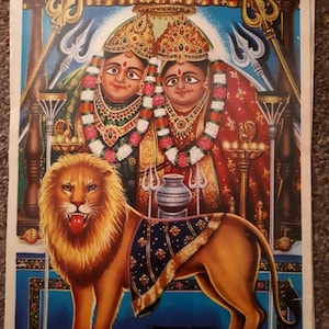 Può includere: Un dipinto religioso colorato che raffigura due figure femminili con elaborate acconciature e gioielli, sedute su un trono con un leone in primo piano. Il dipinto è su sfondo blu con un bordo decorativo. Il testo "Shri Chamunda Mataji Chotila 275" è stampato in basso.