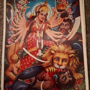 Peut inclure: Une peinture colorée représentant une déesse hindoue aux multiples bras, chevauchant un lion. Elle tue un démon avec une épée. La déesse est ornée de bijoux et de fleurs. La peinture est sur un fond bleu avec une bordure florale.