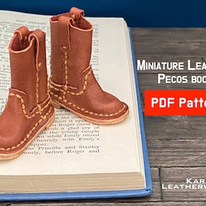 May include: A pair of miniature brown leather cowboy boots with yellow stitching. The boots are sitting on a book with the text "MINIATURE LEATHER PECOS BOOTS PDF Pattern" visible. The text "KARAMEL LEATHERWORK" is also visible.
