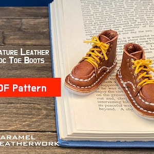 Könnte beinhalten: Ein Paar Miniatur-Stiefeletten aus braunem Leder mit abgerundeter Zehenkappe und gelben Schnürsenkeln. Die Stiefeletten liegen auf einem Buch mit dem Text "MINIATURE LEATHER MOC TOE BOOTS PDF Pattern KARAMEL LEATHERWORK" sichtbar.