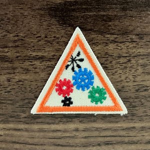 May include: A triangular embroidered patch with an orange border. The patch features a white background with colorful gears and a black star in the center.