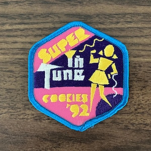 May include: A pink and blue embroidered patch with the text "Super in Tune" and "Cookies '92". A cartoon girl with yellow hair is holding a phone and standing on a purple background.