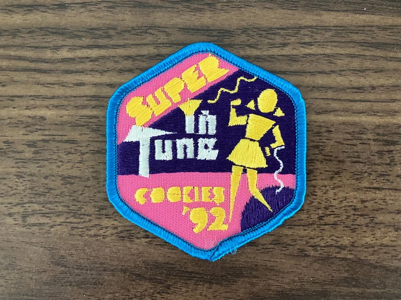 May include: A pink and blue embroidered patch with the text "Super in Tune" and "Cookies '92". A cartoon girl with yellow hair is holding a phone and standing on a purple background.