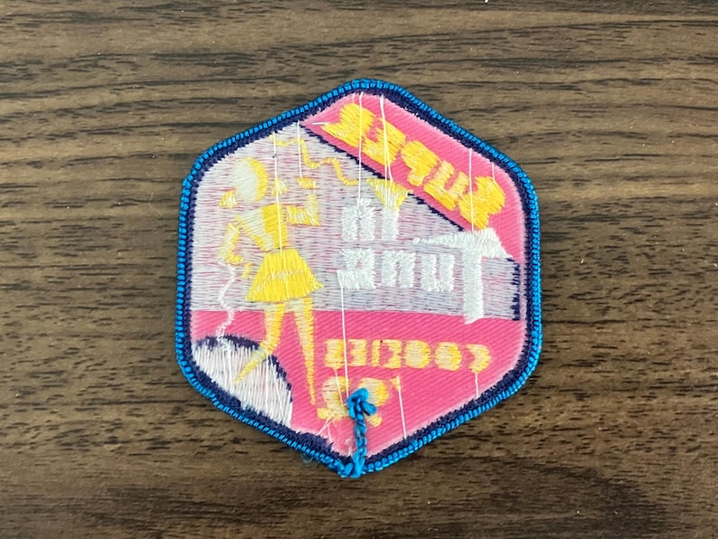 May include: A pink and white embroidered patch with a blue border. The patch features a cartoon girl holding a phone and the text "Betty's Beauty Shop".
