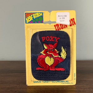 May include: An iron-on patch featuring a red fox with a yellow heart-shaped chest and the word "FOXY" embroidered above. The patch is on a navy blue background and comes in a yellow cardboard package.