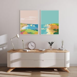 Puede incluir: Dos impresiones enmarcadas de paisajes abstractos coloridos que representan las formaciones rocosas de los Doce Apóstoles en Australia. Las impresiones están colgadas en una pared blanca sobre una cómoda blanca con una tapa de madera.