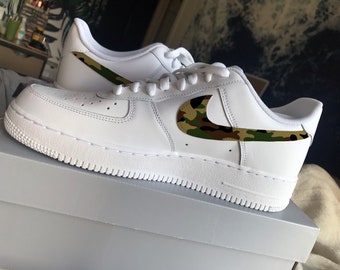 May include: White leather sneakers with a camouflage design on the Nike swoosh and heel tab. The shoes are new and appear to be sitting on a white box. The laces and soles are also white.