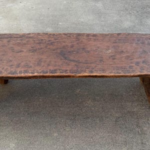 May include: A rustic, rectangular wooden footstool with a dark brown finish. The stool has a slightly curved top and three sturdy, rounded legs. The wood grain is visible, adding to its natural, aged appearance.