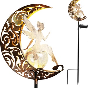 May include: A gold metal fairy sitting on a crescent moon with a solar powered light inside. The fairy is holding a flower and has wings. The moon is decorated with a floral pattern. The solar powered light is on a black metal stake.