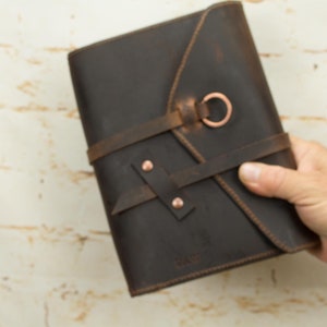 May include: A brown leather journal with a flap closure and a copper ring. The journal has a strap closure with copper rivets and the word "RAW" embossed on the front.