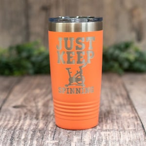 May include: Orange stainless steel tumbler with a silver lid. The tumbler has the text "Just Keep Spinning" and a graphic of a stationary bike.