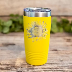 Pode incluir: Um copo de a&ccedil;o inoxid&aacute;vel amarelo com um design de girassol prateado. O copo tem uma tampa e &eacute; isolado.