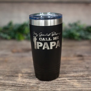 May include: Black stainless steel tumbler with a silver lid. The tumbler has the text "My Greatest Blessings Call Me Papa" engraved on it.
