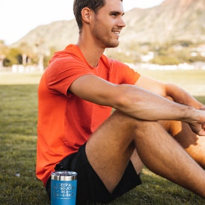 May include: A blue insulated tumbler with the text "I keep all my bad jokes in a dad-a-base" is on the grass. The tumbler has a silver lid. The person in the image is wearing an orange shirt and black shorts.