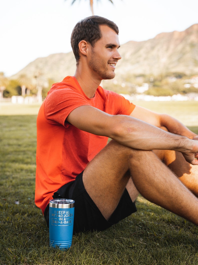 May include: A blue insulated tumbler with the text "I keep all my bad jokes in a dad-a-base" is on the grass. The tumbler has a silver lid. The person in the image is wearing an orange shirt and black shorts.