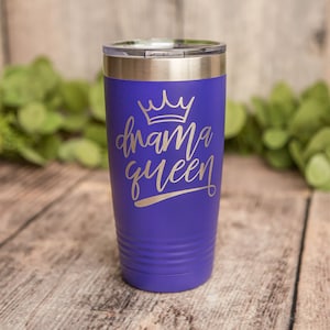 Op de afbeelding: Een paarse roestvrijstalen beker met een zilveren deksel en een gouden kroon en de tekst "drama queen" in een zilveren handschrift.