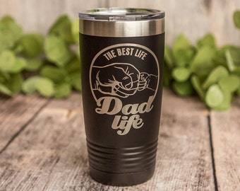 May include: Black stainless steel tumbler with a silver lid. The tumbler has an engraved design of two fists bumping with the text "The Best Life Dad Life" in a circle.