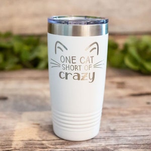 May include: White stainless steel tumbler with a cat face design and the text "One cat short of crazy".