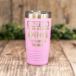 May include: Pink stainless steel tumbler with a gold inscription that reads "Caution I've been known to throw things" with a graphic of four clay pots.