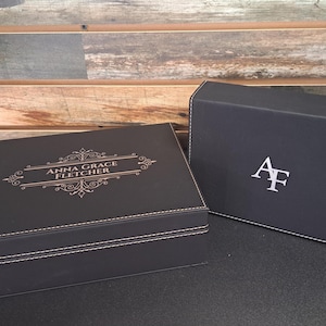 May include: Two black gift boxes with white stitching. The box on the left has the text "Anna Grace Fletcher" in gold lettering. The box on the right has the initials "AF" in white lettering.