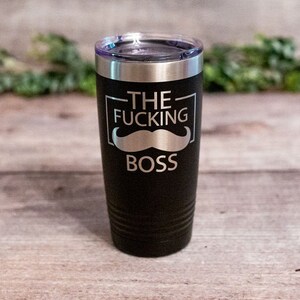May include: Black stainless steel travel mug with a silver inscription that reads "THE FUCKING BOSS" with a silver mustache graphic.