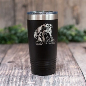 May include: Black insulated tumbler with a silver rim and lid. The tumbler features a detailed etched image of a dog's face. The dog's head and paws are visible, giving the impression it is peeking over the edge.