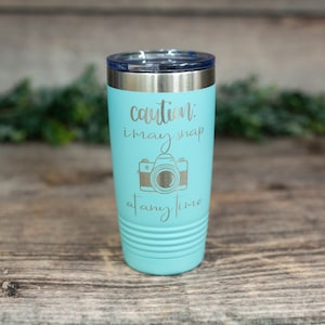 May include: A turquoise stainless steel tumbler with a silver lid and a silver inscription that reads "caution: i may snap at any time" with a camera graphic.